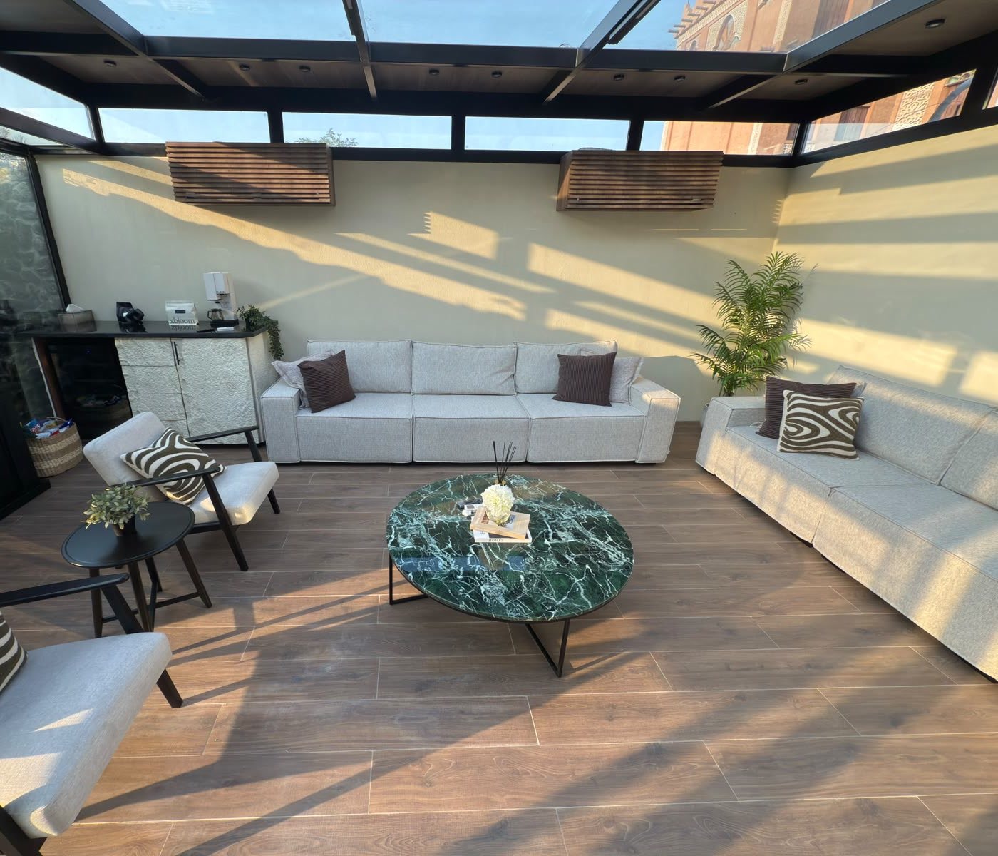 A glass room at golden hour with sofas, a green marble table, and lines of sunlight on the wall